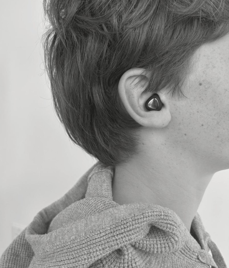 A young man in a grey knit hoodie wearing a hum earplug, shown in muted monochrome